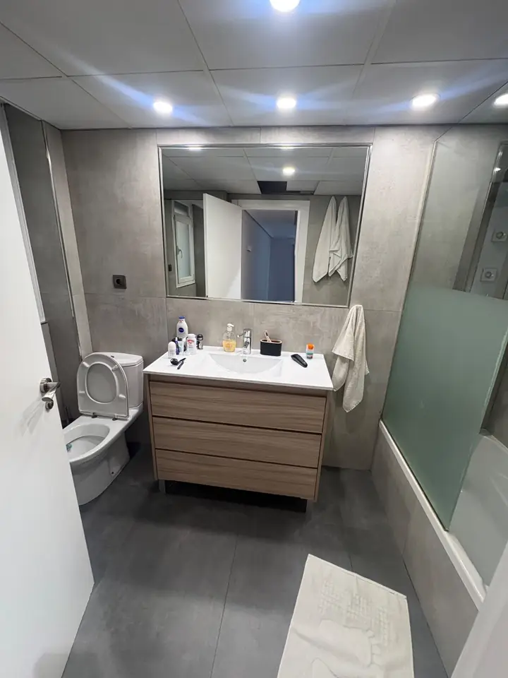 Foto real de un baño antes de la remodelación: lavabo con mueble de madera, inodoro, tina con cancel y azulejo gris